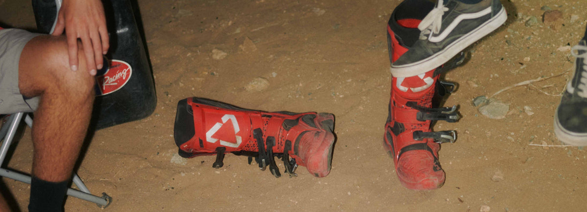 A pair of red and black moto boots with white logo graphics, shown on the ground next to a person sitting on a folding chair in a dirt setting.