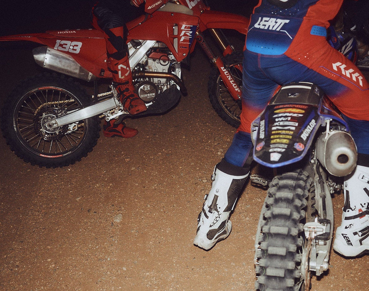 A motocross rider in red, blue, and white Leatt gear sits on a dirt bike at night on a dirt track, with other bikes partially visible in the background.